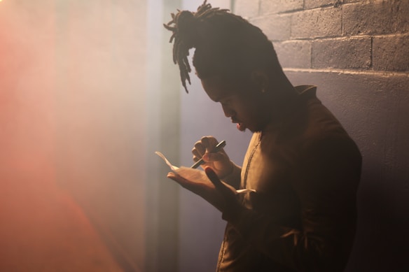 A person is standing against a brick wall, focusing intently on writing in a notebook held in their hand. The scene is dimly lit, with warm light creating soft shadows and a contemplative atmosphere.