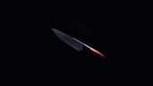 A sharp kitchen knife with a metallic blade and a red handle is illuminated against a dark background, emphasizing its sleek and shiny appearance.