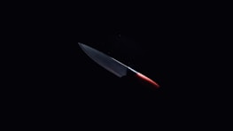 A sharp kitchen knife with a metallic blade and a red handle is illuminated against a dark background, emphasizing its sleek and shiny appearance.