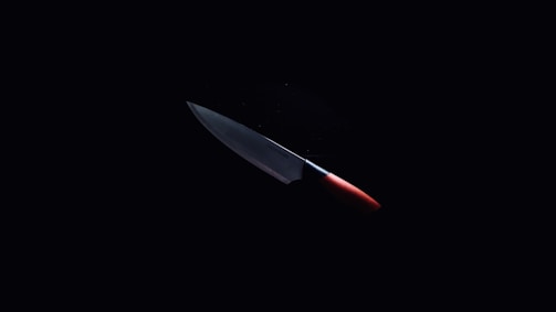 A sharp kitchen knife with a metallic blade and a red handle is illuminated against a dark background, emphasizing its sleek and shiny appearance.