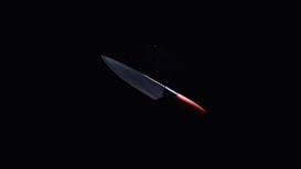 A sharp kitchen knife with a metallic blade and a red handle is illuminated against a dark background, emphasizing its sleek and shiny appearance.