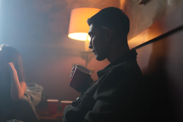 Illustration of a grumpy older man holding a coffee cup in a dimly lit recovery meeting room with folding chairs.