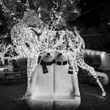 Two polar bear sculptures wear scarves and sit closely together, forming a whimsical bench. Behind them is a large, illuminated wire-frame sculpture of a deer adorned with sparkling lights. The background features trees decorated with strings of lights, enhancing the festive atmosphere.