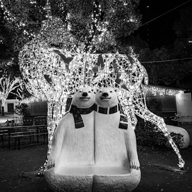 Two polar bear sculptures wear scarves and sit closely together, forming a whimsical bench. Behind them is a large, illuminated wire-frame sculpture of a deer adorned with sparkling lights. The background features trees decorated with strings of lights, enhancing the festive atmosphere.