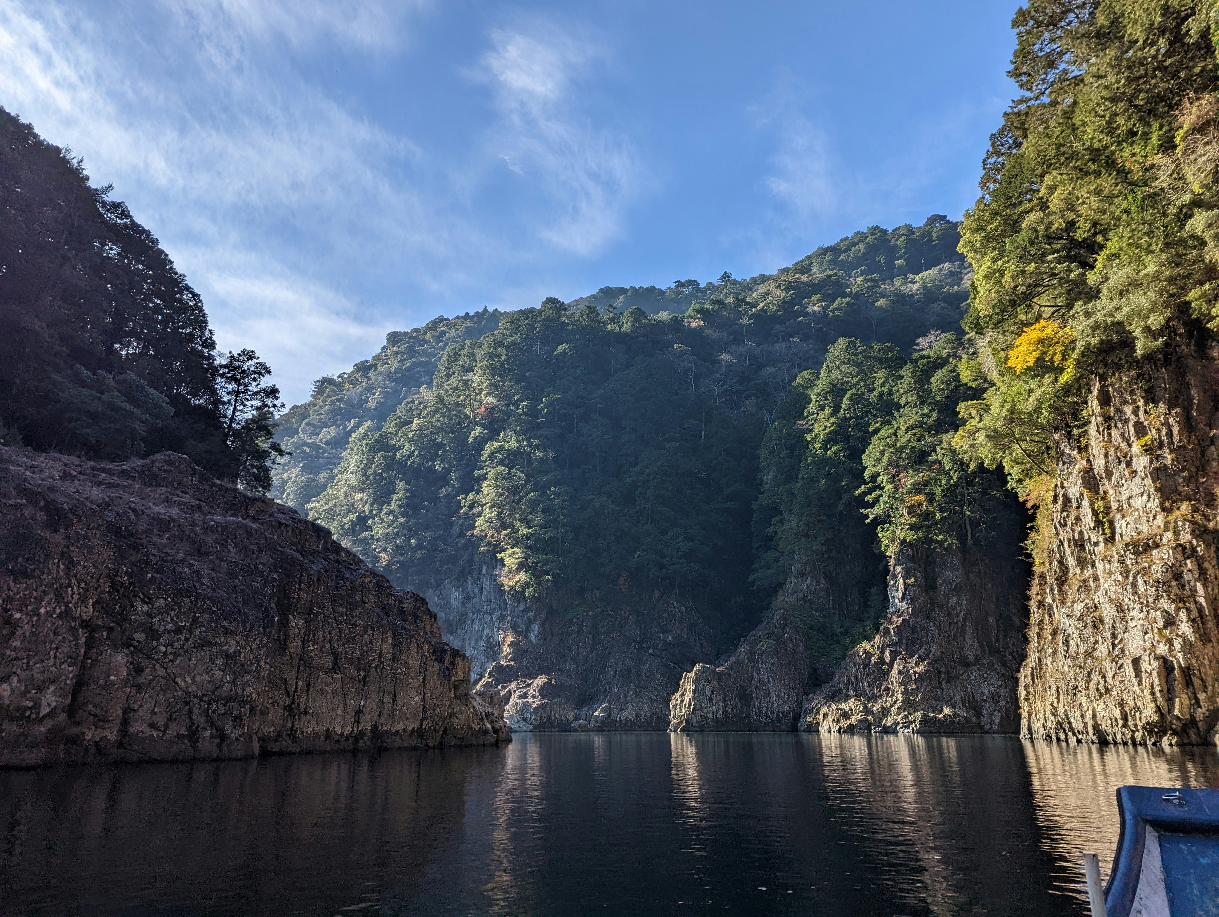 a boat is in the water near a mountain, The Doro-kyo Valley in Japan is an incredible sight, with its huge cliffs, tall trees, and serene mountain reflections in the river below. The valley is so large and peaceful, covered in rich greenery - it