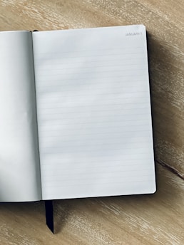 An open notebook is placed on a wooden surface, with its pages displaying fine blue lines for writing. In the top-right corner of the page, the date 'January 1' is printed.