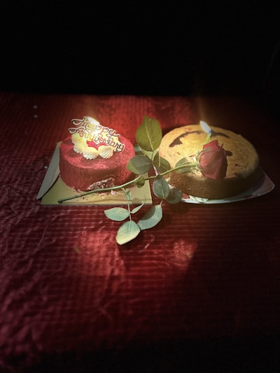 Two cakes, one red velvet with a 'Happy Anniversary' topper and the other a plain cake, are placed on a red textured surface. A lit candle and a single red rose with green leaves are present, adding to the celebratory mood.