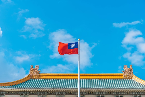 A flag with a red, white, and blue design waves prominently against a clear blue sky. It is mounted on a tall pole above a decorative building roof featuring ornate details and vibrant colors. The architecture appears traditional with intricate carvings and a mix of teal, yellow, and gold hues.