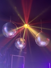 Three disco balls are suspended from the ceiling, with colorful lights reflecting off their surfaces. A central light source emits red, orange, and yellow beams spreading across the dark background, creating a vibrant and festive atmosphere.