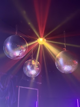 Three disco balls are suspended from the ceiling, with colorful lights reflecting off their surfaces. A central light source emits red, orange, and yellow beams spreading across the dark background, creating a vibrant and festive atmosphere.