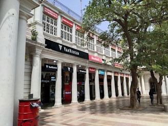 A series of commercial storefronts with white columns and large glass windows, located on the ground floor of a classical-style building. The shops include known brands like Van Heusen and others. Above the stores, there are more windows with red signs and an upper balcony. There is a tree with green foliage on the right side, and several people can be seen walking or standing around the area. A red postbox with 'LETTERS' written on it is visible in the foreground on the left side.