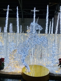 A wireframe sculpture of a unicorn illuminated with white and blue lights stands on a golden circular platform. Surrounding the unicorn are vertical light structures resembling icicles, with additional tree-shaped light decorations. The scene has a festive and enchanting ambiance, accented with nearby red poinsettias.