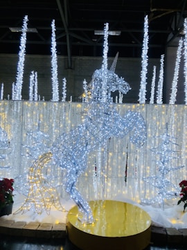 A wireframe sculpture of a unicorn illuminated with white and blue lights stands on a golden circular platform. Surrounding the unicorn are vertical light structures resembling icicles, with additional tree-shaped light decorations. The scene has a festive and enchanting ambiance, accented with nearby red poinsettias.