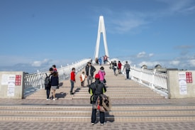 A bustling pedestrian bridge with multiple people walking across it, featuring a prominent white arch structure. The bridge is flanked by railings, and there are signs in a foreign language. The sky is clear with a few clouds, suggesting a bright and sunny day.