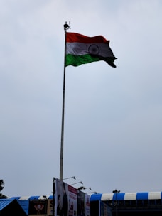 A tall flagpole displaying the Indian national flag against a cloudy sky. The area below shows some rooftops with blue accents and a few billboards.