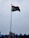 A tall flagpole displaying the Indian national flag against a cloudy sky. The area below shows some rooftops with blue accents and a few billboards.