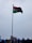 A tall flagpole displaying the Indian national flag against a cloudy sky. The area below shows some rooftops with blue accents and a few billboards.