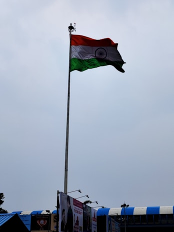 A tall flagpole displaying the Indian national flag against a cloudy sky. The area below shows some rooftops with blue accents and a few billboards.