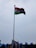 A tall flagpole displaying the Indian national flag against a cloudy sky. The area below shows some rooftops with blue accents and a few billboards.