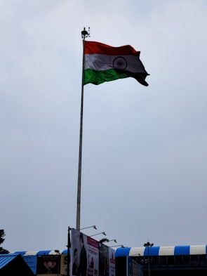 A tall flagpole displaying the Indian national flag against a cloudy sky. The area below shows some rooftops with blue accents and a few billboards.
