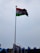 A tall flagpole displaying the Indian national flag against a cloudy sky. The area below shows some rooftops with blue accents and a few billboards.