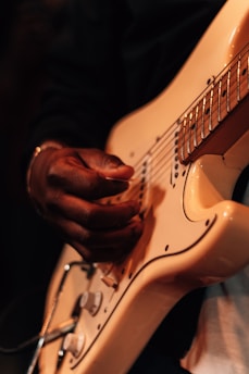 A close-up of a person's hand playing a beige electric guitar. The fingers are positioned on the strings, and the lighting gives a warm, intimate feel to the scene.