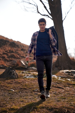 A person is walking through a natural, outdoor setting, holding a jacket over their shoulder and sunglasses in one hand. They are dressed in a casual outfit consisting of a plaid shirt, dark jeans, and sneakers. The background features leafless trees and earthy ground, creating an autumnal or early spring atmosphere.