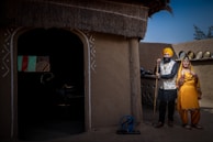 A man and woman in traditional attire are standing outside an earthen-walled building with a thatched roof. The man is wearing a turban and holding a stick, while the woman is dressed in a vibrant yellow outfit with colorful embellishments and jewelry. The background features traditional elements like an old spinning wheel and metal pots arranged neatly on a ledge.