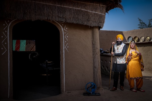 A man and woman in traditional attire are standing outside an earthen-walled building with a thatched roof. The man is wearing a turban and holding a stick, while the woman is dressed in a vibrant yellow outfit with colorful embellishments and jewelry. The background features traditional elements like an old spinning wheel and metal pots arranged neatly on a ledge.