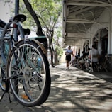 A city street scene featuring a bicycle parked near a lamp post surrounded by green trees. People are sitting and conversing at an outdoor cafe with a sign reading 'Little French'. A person appears to be walking and holding something along the sidewalk which is lined with cobblestones.