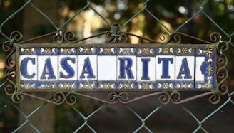 A decorative ceramic tile sign mounted on a wire fence, framed with ornate wrought iron swirls, displaying the words 'CASA RITA' in blue letters with floral patterns in the background. The setting appears to be outdoors with a blurred natural backdrop.