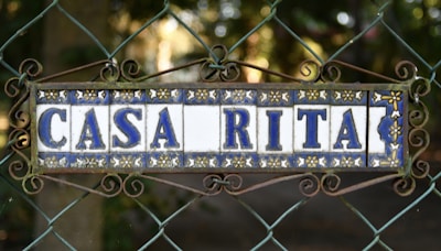 A decorative ceramic tile sign mounted on a wire fence, framed with ornate wrought iron swirls, displaying the words 'CASA RITA' in blue letters with floral patterns in the background. The setting appears to be outdoors with a blurred natural backdrop.