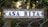 A decorative ceramic tile sign mounted on a wire fence, framed with ornate wrought iron swirls, displaying the words 'CASA RITA' in blue letters with floral patterns in the background. The setting appears to be outdoors with a blurred natural backdrop.