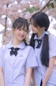 Two young people in school uniforms are standing together in front of cherry blossoms. They wear matching light blue shirts with black bow ties. One person is smiling at the camera while the other looks affectionately at them.