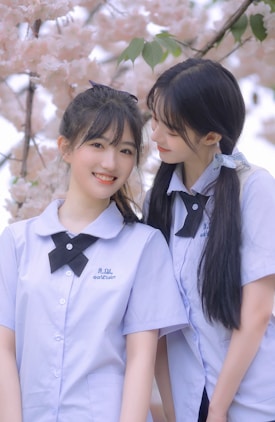 Two young people in school uniforms are standing together in front of cherry blossoms. They wear matching light blue shirts with black bow ties. One person is smiling at the camera while the other looks affectionately at them.