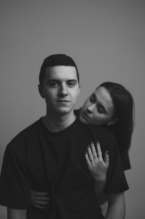 A black and white photograph showing a man in a dark shirt looking directly at the camera with a neutral expression. Behind him, a woman with long hair leans on his back with her head resting on his shoulder, eyes closed, and her arms wrapped around him. The background is plain, adding focus to the subjects.