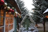 Snow-covered Christmas decorations adorn small wooden huts, with garlands of colorful ornaments hanging from the eaves. Tall evergreen trees dusted with snow flank the scene, creating a festive winter atmosphere.