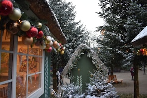 Snow-covered Christmas decorations adorn small wooden huts, with garlands of colorful ornaments hanging from the eaves. Tall evergreen trees dusted with snow flank the scene, creating a festive winter atmosphere.