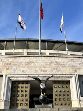 A large building with multiple flagpoles and flags positioned above a wide entrance. A prominent statue of an eagle stands in front of the entrance, mounted on a spherical base. The building has a detailed stone facade and the sky is clear.