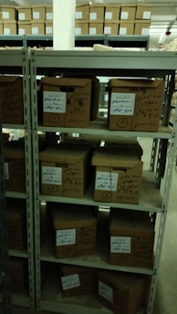 A set of metal shelves filled with cardboard boxes, each labeled with handwritten notes. The shelves are located in a storage area, possibly a warehouse or archive, with additional boxes in the background. The lighting is dim, creating a somewhat shadowy environment.