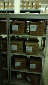 A set of metal shelves filled with cardboard boxes, each labeled with handwritten notes. The shelves are located in a storage area, possibly a warehouse or archive, with additional boxes in the background. The lighting is dim, creating a somewhat shadowy environment.