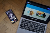 A smartphone and a laptop are placed on a wooden floor. The smartphone displays a dark-themed social networking app with the text 'Social networking that's not for sale.' The laptop screen shows a colorful cartoon illustration with animated animals, including what appears to be a mammoth, in a welcoming scene.