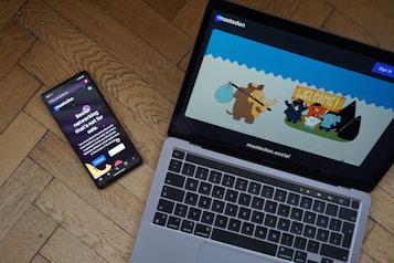A smartphone and a laptop are placed on a wooden floor. The smartphone displays a dark-themed social networking app with the text 'Social networking that's not for sale.' The laptop screen shows a colorful cartoon illustration with animated animals, including what appears to be a mammoth, in a welcoming scene.
