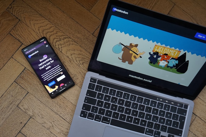 A smartphone and a laptop are placed on a wooden floor. The smartphone displays a dark-themed social networking app with the text 'Social networking that's not for sale.' The laptop screen shows a colorful cartoon illustration with animated animals, including what appears to be a mammoth, in a welcoming scene.