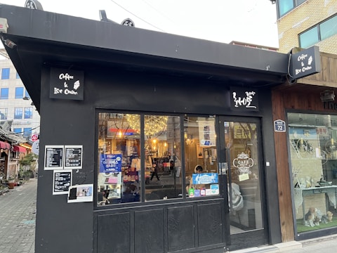 A small black cafe with signs advertising coffee and ice cream. The entrance features glass doors with interior lights visible inside. A board displaying a menu is attached to the wall. The surroundings include a cozy street with a few potted plants and neighboring buildings.