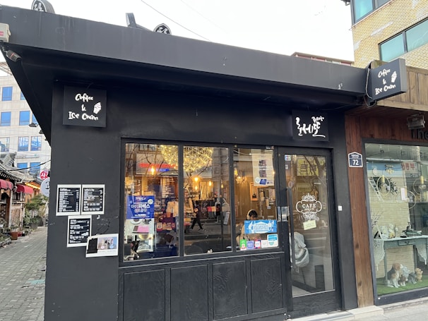 A small black cafe with signs advertising coffee and ice cream. The entrance features glass doors with interior lights visible inside. A board displaying a menu is attached to the wall. The surroundings include a cozy street with a few potted plants and neighboring buildings.