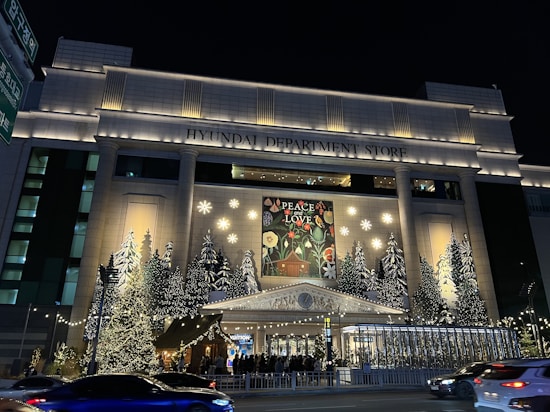 A large department store is beautifully decorated with numerous Christmas trees adorned with lights. A prominent sign above the entrance reads 'Peace and Love' surrounded by floral patterns. The facade features elegant architectural elements such as columns and a pediment. The night setting enhances the glow of the lights, creating a festive and inviting atmosphere.