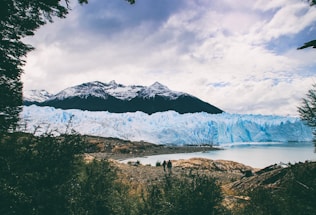 A breathtaking view featuring towering mountains with snow-capped peaks in the background. A vast, glistening glacier stretches across the scene, its icy blue hues contrasting with the rugged terrain. Two small figures stand by a lake in the foreground, framed by dense greenery and rocks, all under a partly cloudy sky.