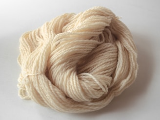 A neatly coiled skein of cream-colored yarn, displaying a soft texture and tightly wound appearance. The yarn has a natural, unbleached look and is resting on a pale surface.