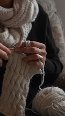 A person is knitting with beige yarn, creating a textured, patterned cloth. They are wearing a cozy scarf and a dark sweater. The focus is on their hands, which hold knitting needles and the partially completed knitting. A ball of yarn and their rings are also visible.